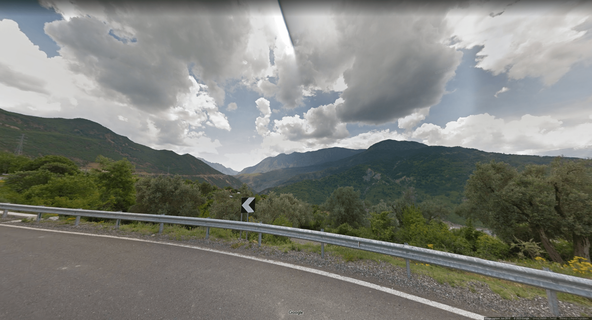 Picture of a beautiful valley area with hills and mountains, and a white on black turn chevron on the road